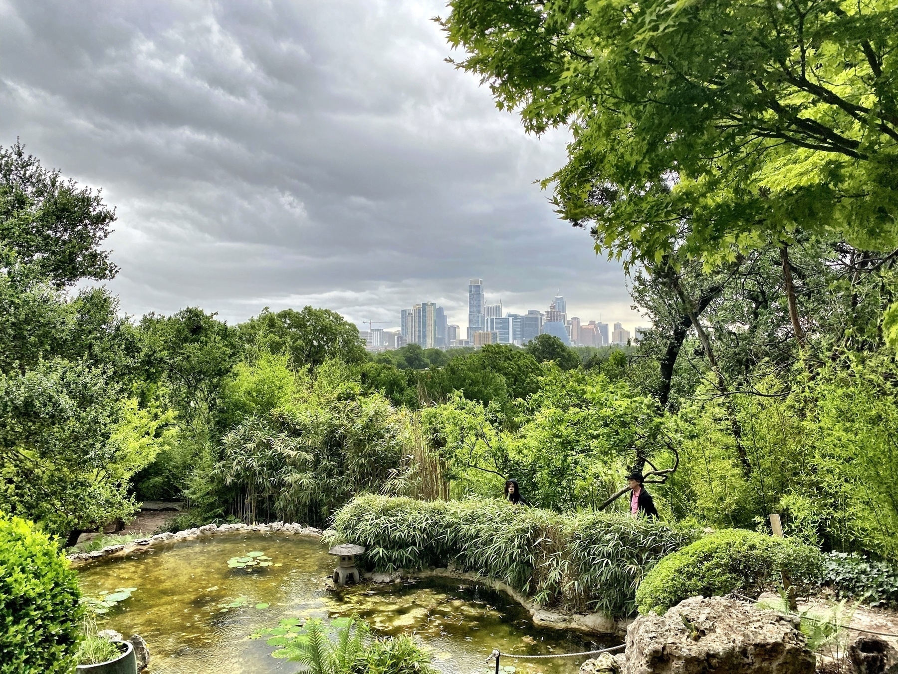 A lush green garden with a pond and city skyline in the background is shown under a cloudy sky.