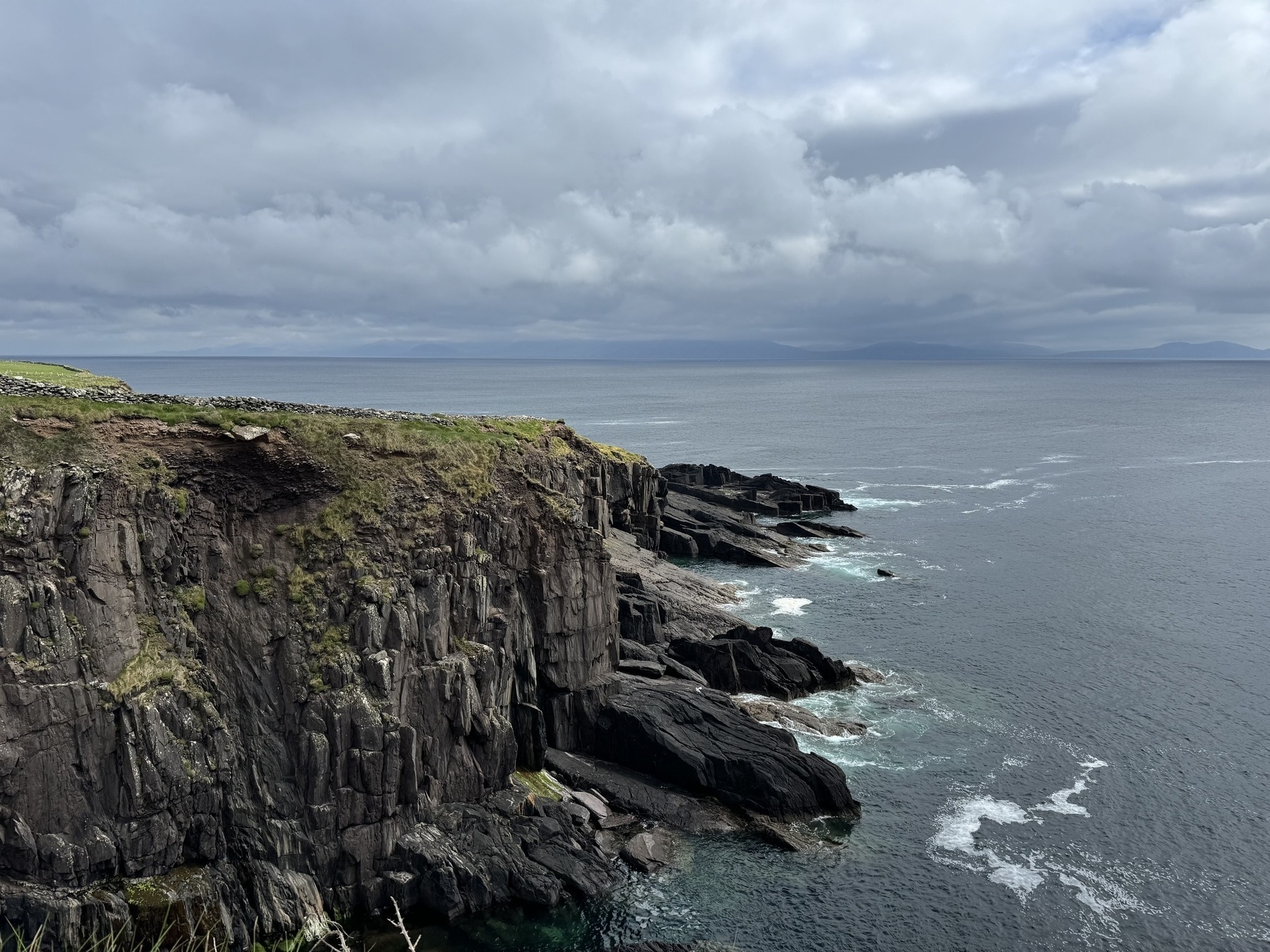 A rugged coastline with rocky cliffs overlooks a vast, cloudy ocean under a slightly overcast sky.