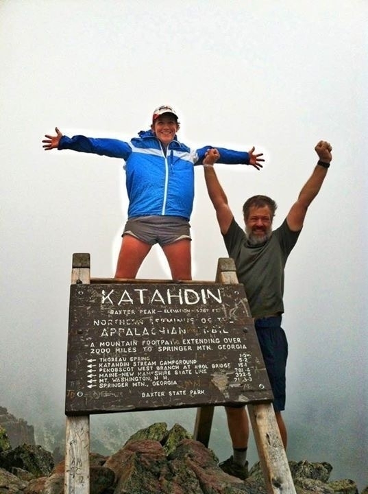 Two people celebrating on top of a mountain summit beside the Katahdin sign, indicating the northern terminus of the Appalachian Trail.