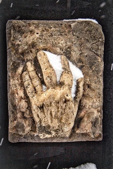  A textured stone relief sculpture depicting a hand with extended three fingers and thumb, partially covered with snow. The high-relief carving is set against a rough, rectangular, stone-like background, with some smaller patches of snow around the edges.