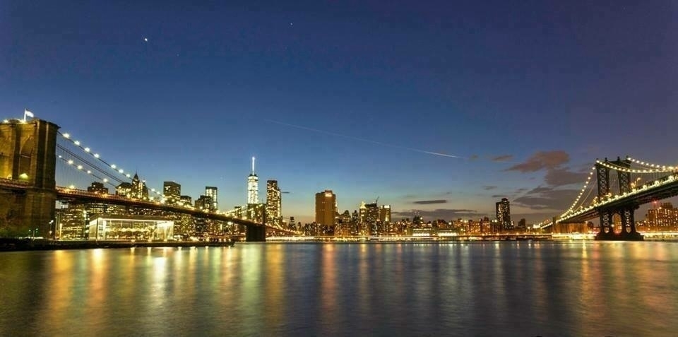 A skyline view featuring two illuminated bridges and a cityscape reflected in the calm waters at dusk.