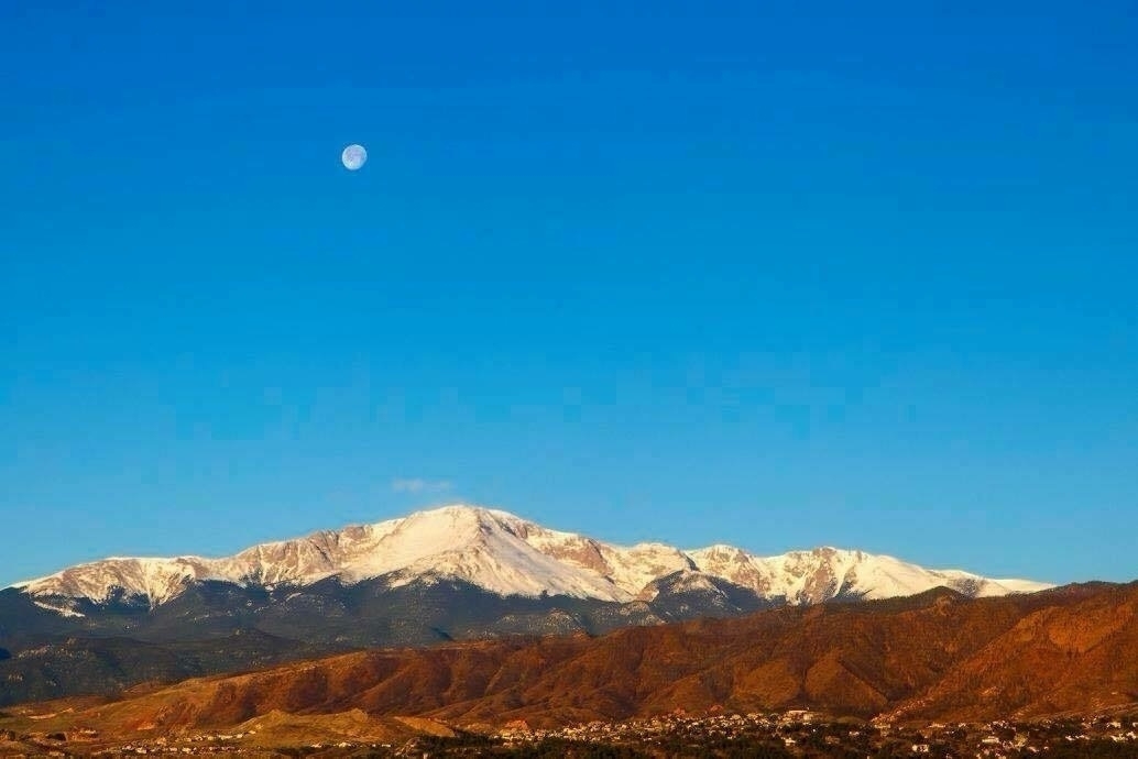 The image shows a clear blue sky with the moon visible in the upper left. Below, a range of snow-capped mountains stretches across the horizon. In the foreground, there are smaller hills and a few scattered buildings, with a landscape that transitions from green to brown.