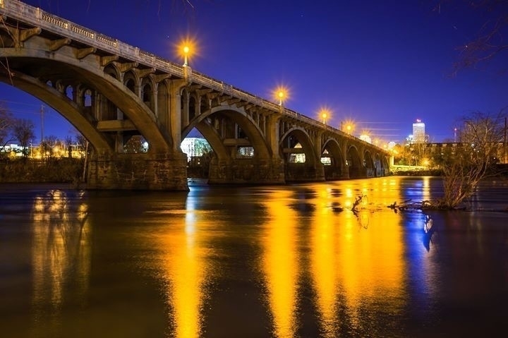 A bridge with multiple arches is illuminated by streetlights at night, casting bright yellow reflections on the calm water below. In the background, city buildings with lights on are faintly visible under a deep blue sky. Bare branches are partially seen on the right and left edges of the image.