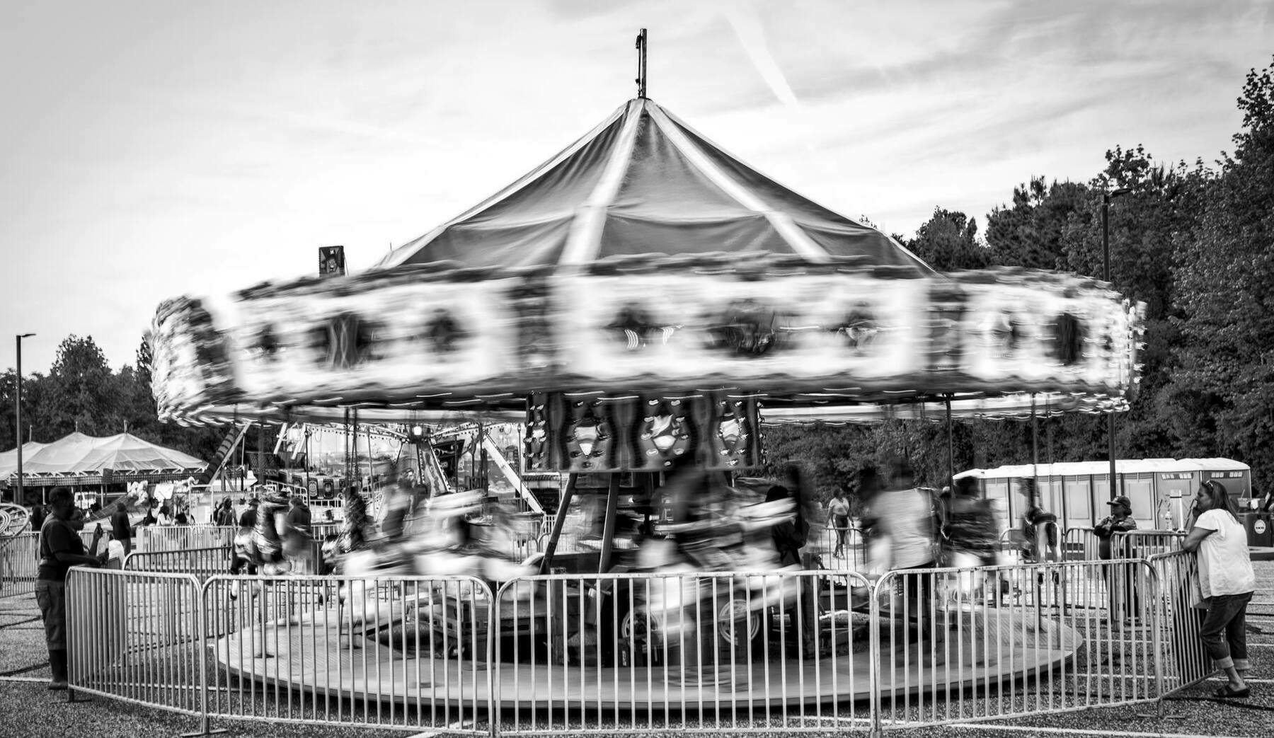 A black and white image of a spinning carousel at a fairground. The motion blur of the carousel creates a dynamic effect, capturing the movement of the ride. The carousel is surrounded by a metal fence and several people, including adults and children, are visible near the ride, with some watching it in motion. Trees and tents can be seen in the background under a partly cloudy sky.