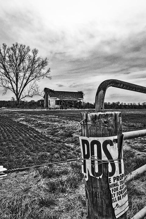 Black and white photo of a Posted sign on the gate into a field where a white wooden farm house sits