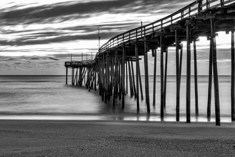 A black and white image of a long wooden pier extending over calm water. The pier's supports cast reflections on the water, creating a serene atmosphere. Above, the sky is filled with streaks of clouds, adding texture to the scene. The shoreline is visible in the foreground, with a smooth, sandy surface.