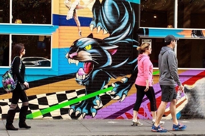 Colorful street art featuring a fierce black panther with sharp claws and an intense gaze is painted on a brick wall. Three people are walking past it on the sidewalk; they appear to be engaged in casual activity. The mural is vibrant with shades of blue, pink, and green, adding a dynamic backdrop to the scene.