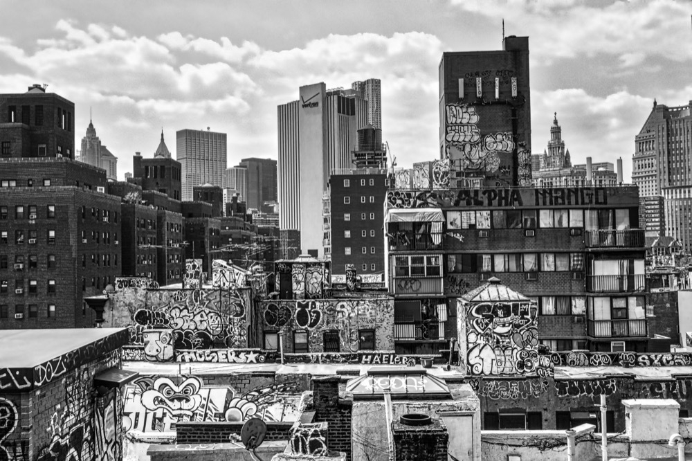 Black and white photo showcasing an urban cityscape with graffiti-covered rooftops in the foreground. High-rise buildings and skyscrapers form the backdrop under a partly cloudy sky. The buildings are adorned with various graffiti tags and artwork, contributing to the gritty, artistic atmosphere of the scene.