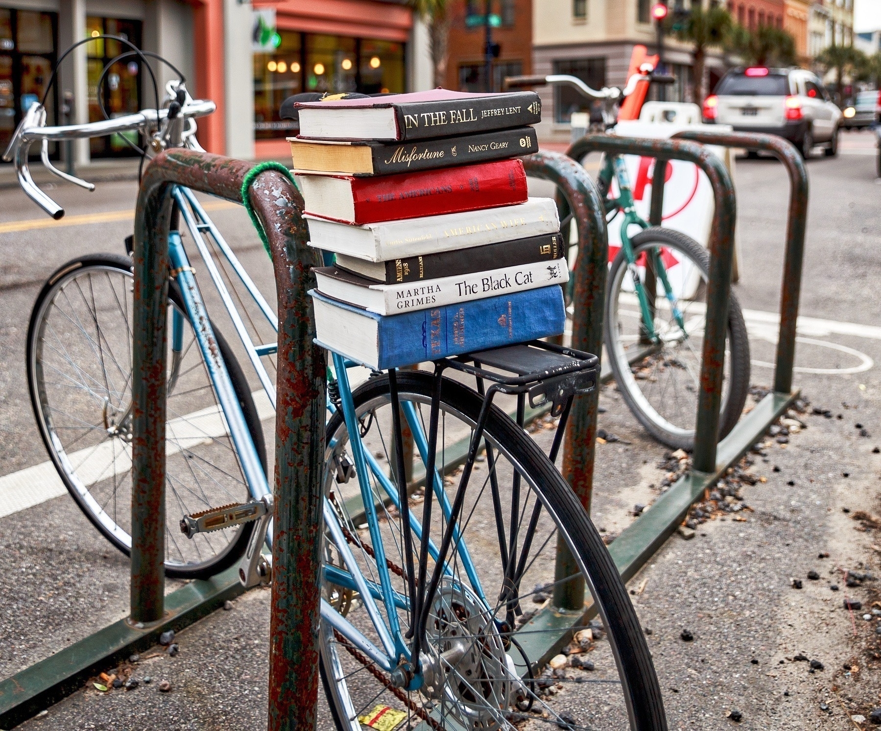 A stack of books is placed creatively on the seat of a bicycle parked at a metal bike rack on a city street.