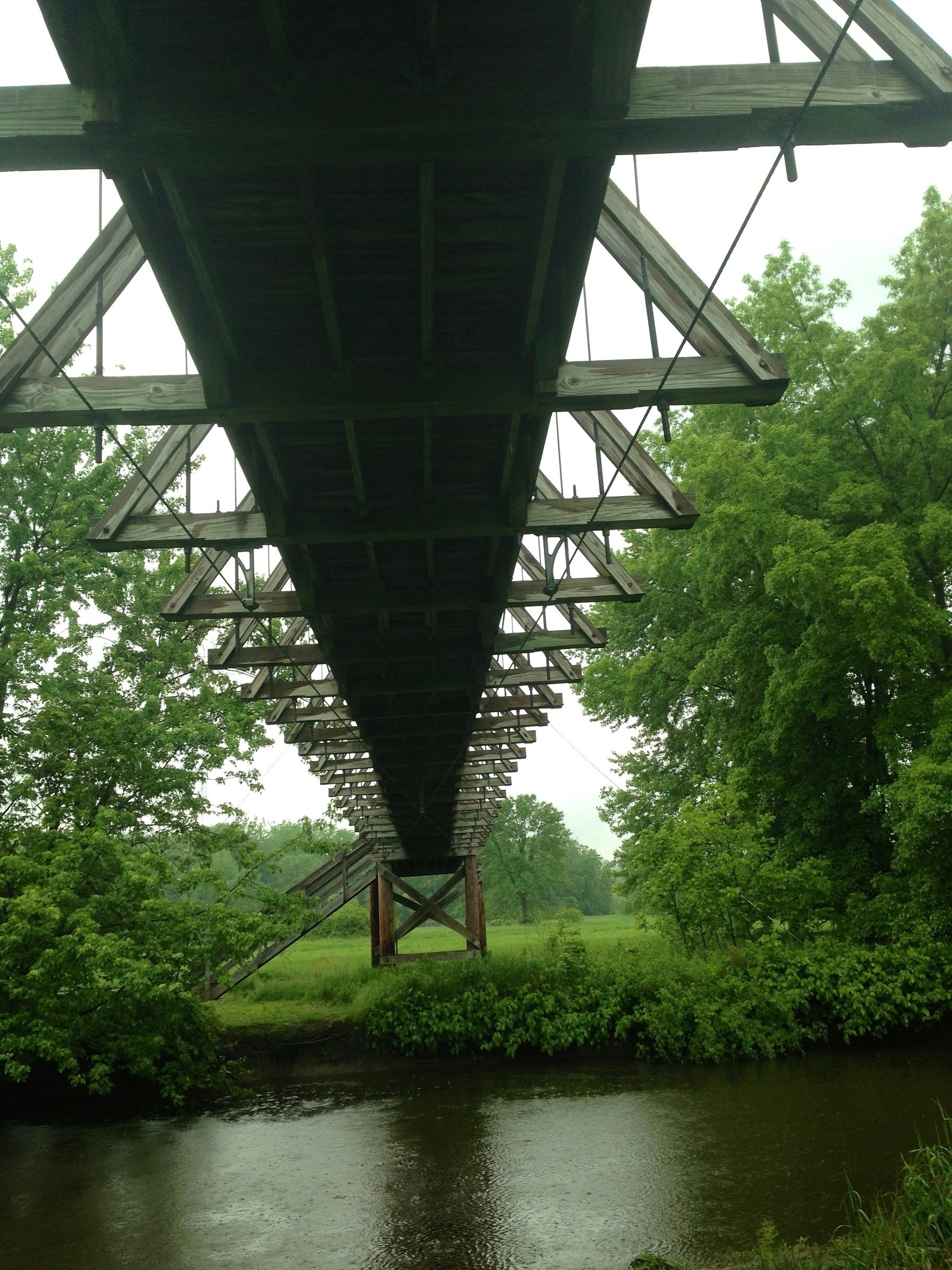 A wooden bridge with a triangular truss design spans over a river, surrounded by lush green trees.