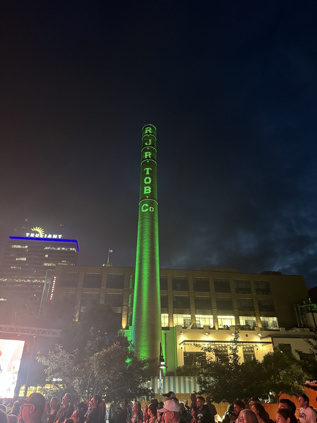 夜晚，建筑物前矗立着一座高大的烟囱或塔楼，灯火通明。结构上可见字母“CAPCO”。周围地区挤满了人，可能是在参加活动。背景是部分多云的夜空。