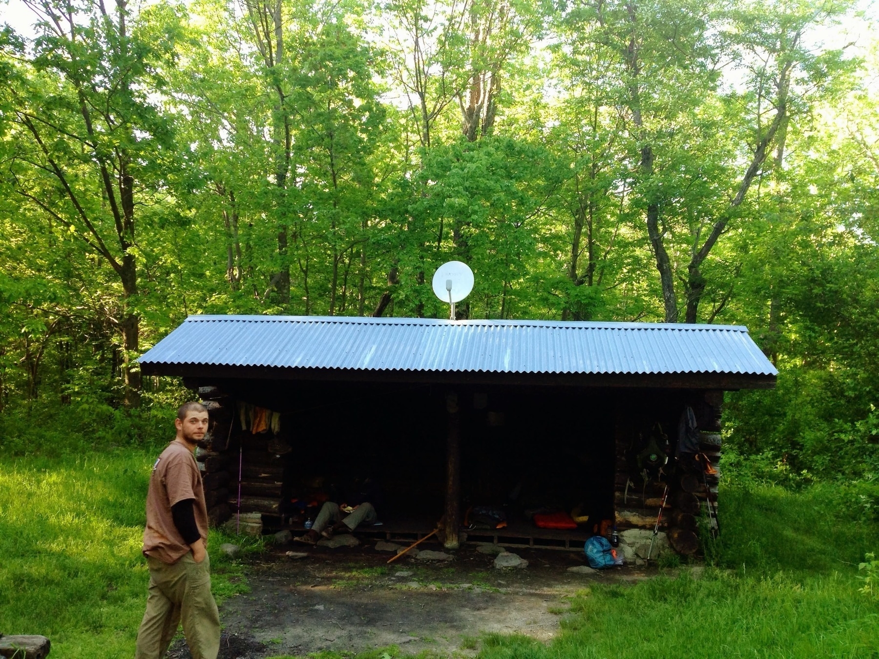 A person is walking near a small wooden shelter in a forest. The shelter has a corrugated metal roof and a satellite dish on top. The surrounding area is lush with green trees, and there are backpacks and gear visible inside the shelter. The ground around the shelter is grassy with some dirt patches.