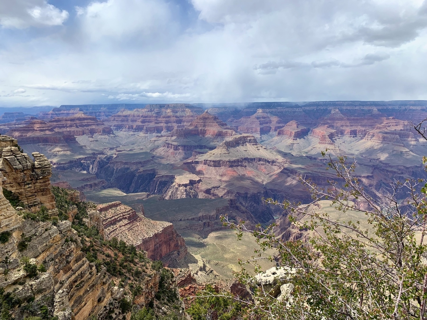 A breathtaking view of the Grand Canyon, showcasing its vast, rugged landscape with multi-layered rock formations in shades of red, orange, and brown. The canyon stretches into the distance under a partly cloudy sky, with shadows cast across the terrain. In the foreground, a rocky ledge with some greenery is visible.