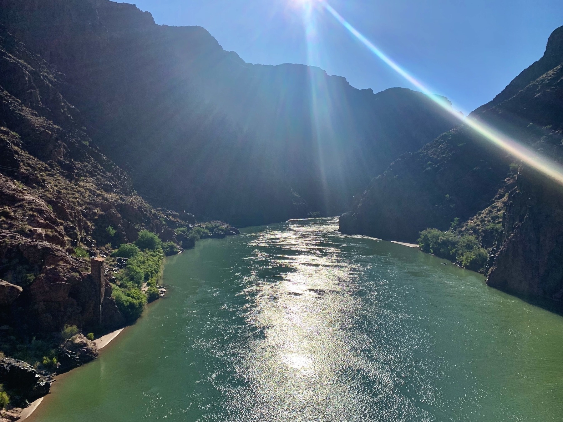 A river flowing through a rocky canyon with steep cliffs on either side. The sunlight casts bright rays across the sky, illuminating the water's surface and creating a shimmering effect. Sparse greenery clings to the rugged terrain, adding patches of vibrant color to the landscape.
