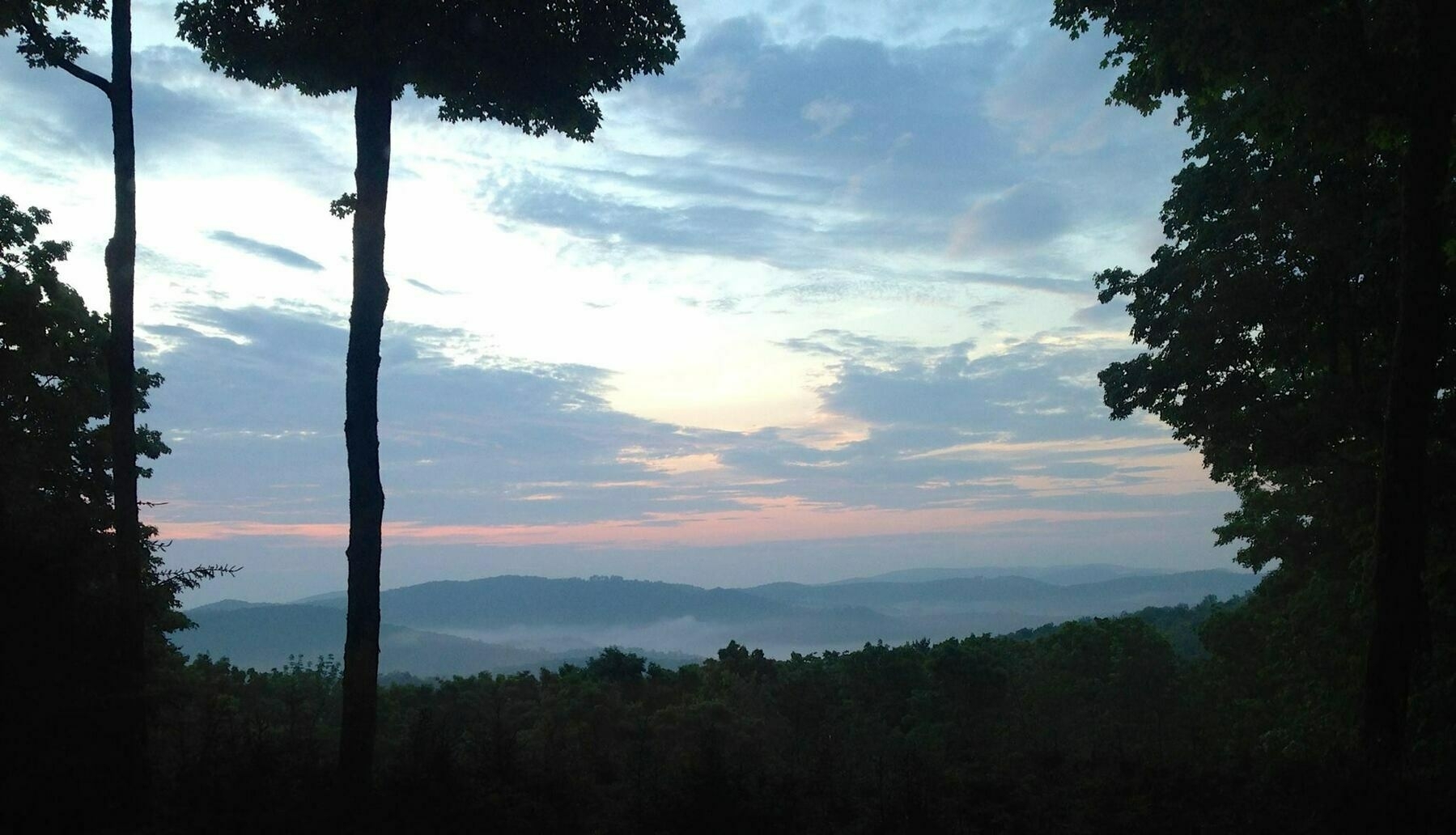 A scenic view of a mountain range at sunrise. The sky is a mix of blues, whites, and pinks/oranges, suggesting the transition between day and night. There's a layer of fog or mist hanging over the lower parts of the mountains, giving them a hazy, layered appearance. The foreground is framed by dark silhouettes of trees on either side and in the center, creating a natural frame for the landscape. The overall impression is one of tranquility and natural beauty.