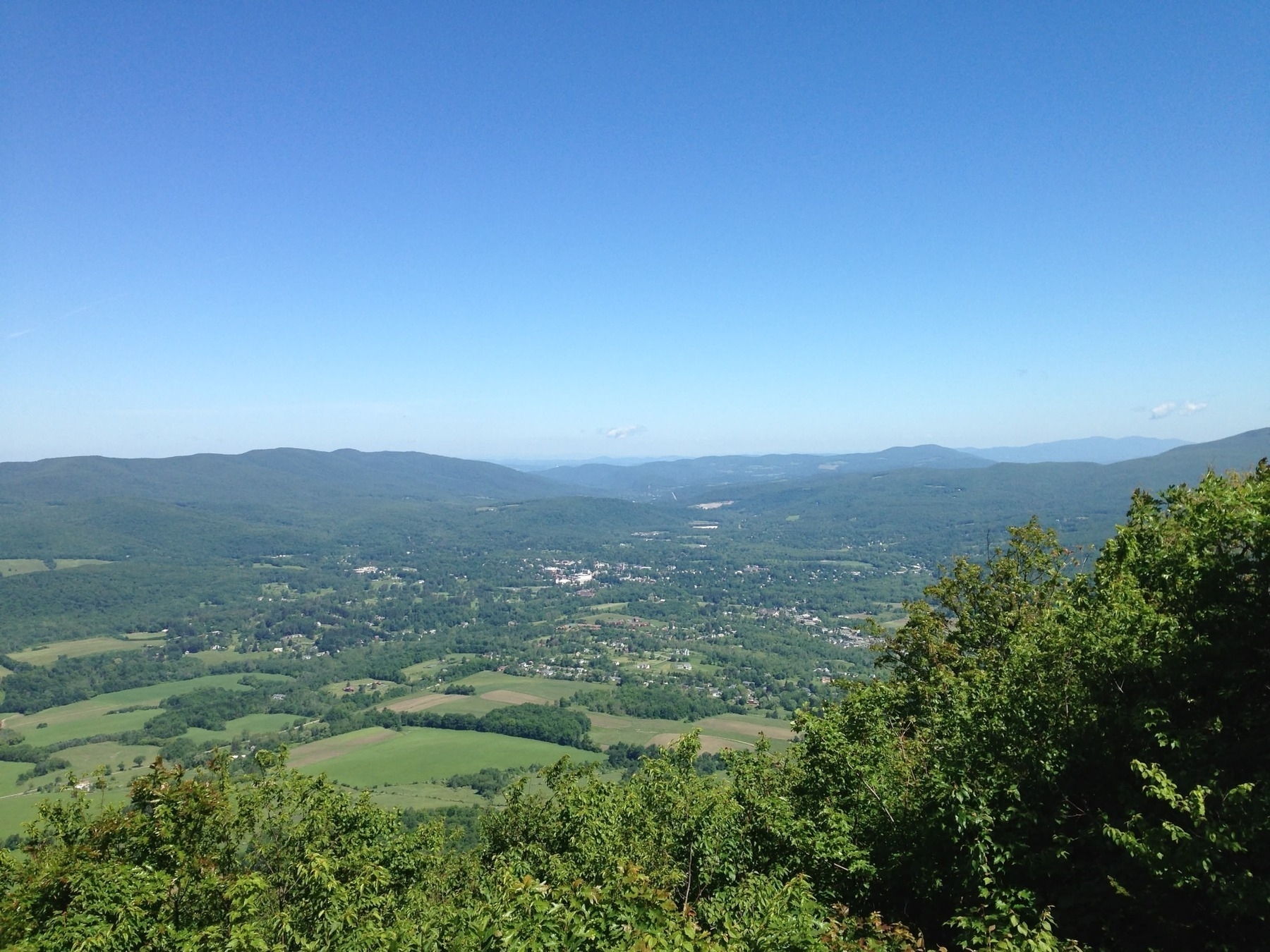 A scenic view of a lush green valley with rolling hills in the background, under a clear blue sky. The valley is dotted with patches of farmland and clusters of trees, and there are small towns scattered throughout the landscape. Dense green foliage is visible in the foreground.
