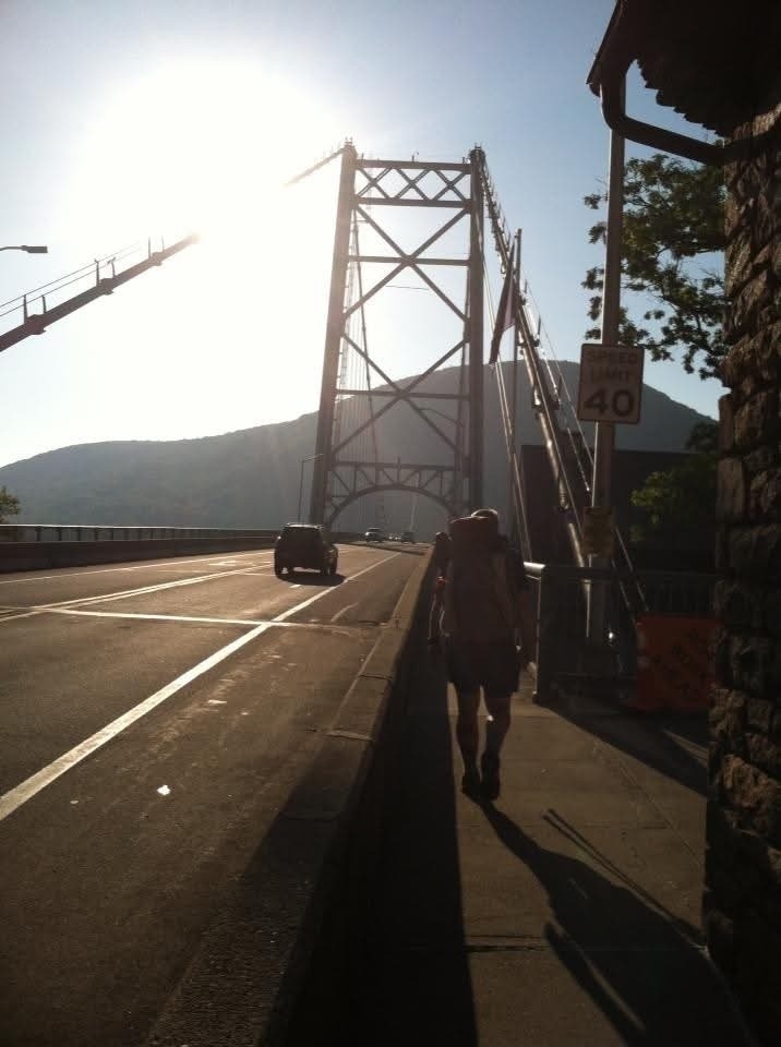 A person is walking along the side of a road on a bridge with a mountain in the background and a vehicle approaching.