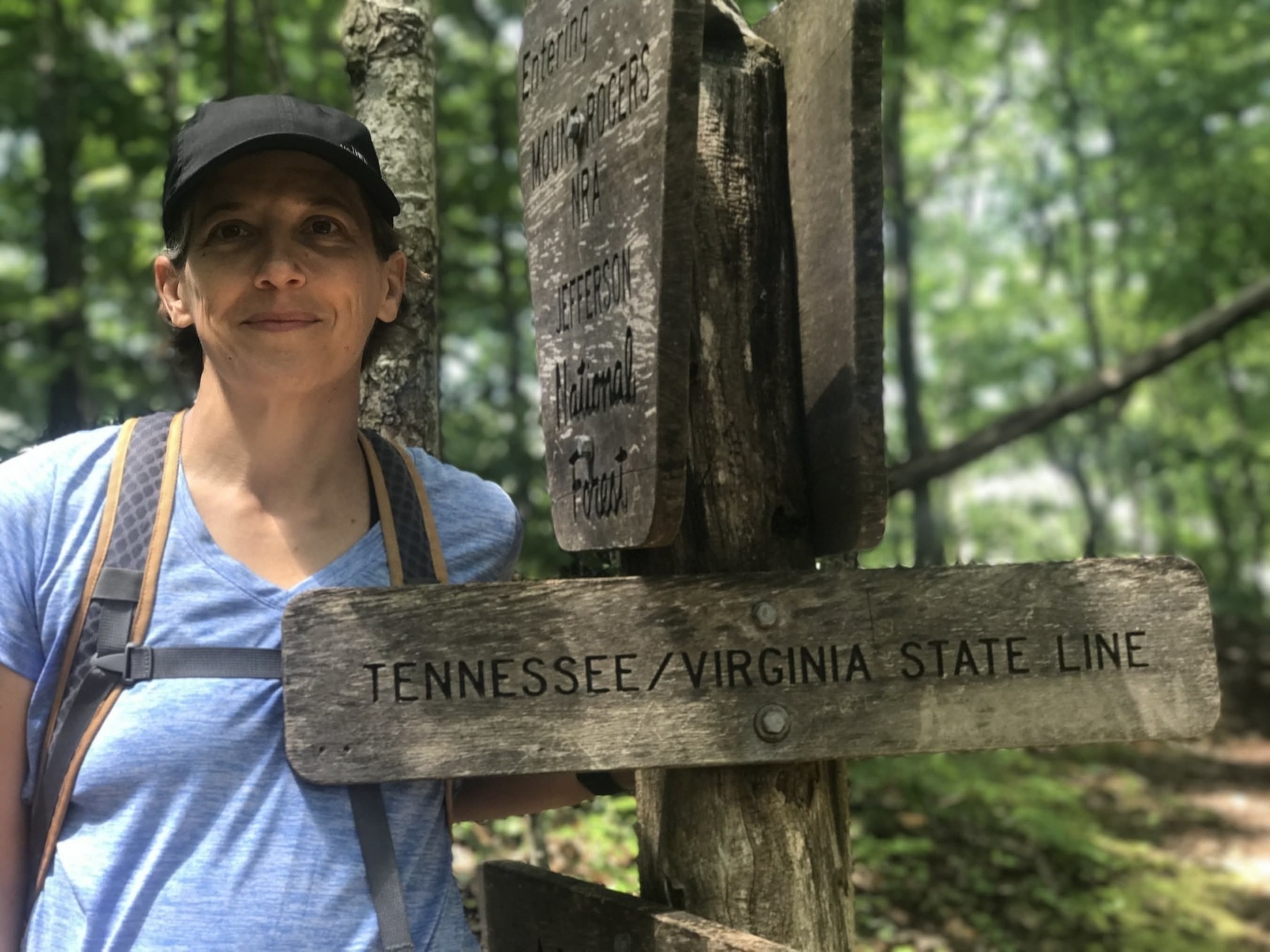 A person wearing a blue shirt and black cap stands beside a wooden sign in the forest. The sign reads "Tennessee/Virginia State Line." The background features lush green trees.