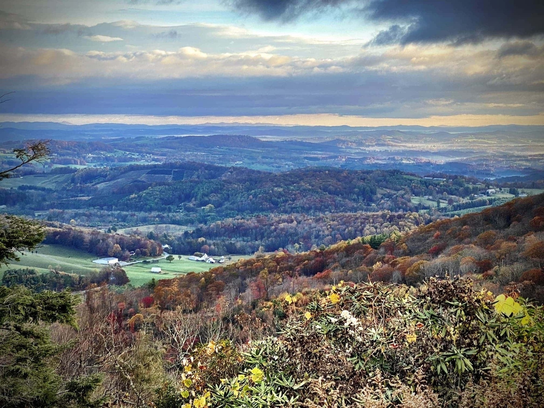 A scenic landscape of rolling hills covered in autumn foliage. The foreground features dense shrubs with yellow and orange leaves. In the distance, the hills display a mix of green, orange, and brown trees, under a sky filled with dramatic clouds. A few scattered farm buildings are visible across the fields and valleys, adding a rural touch to the scene.