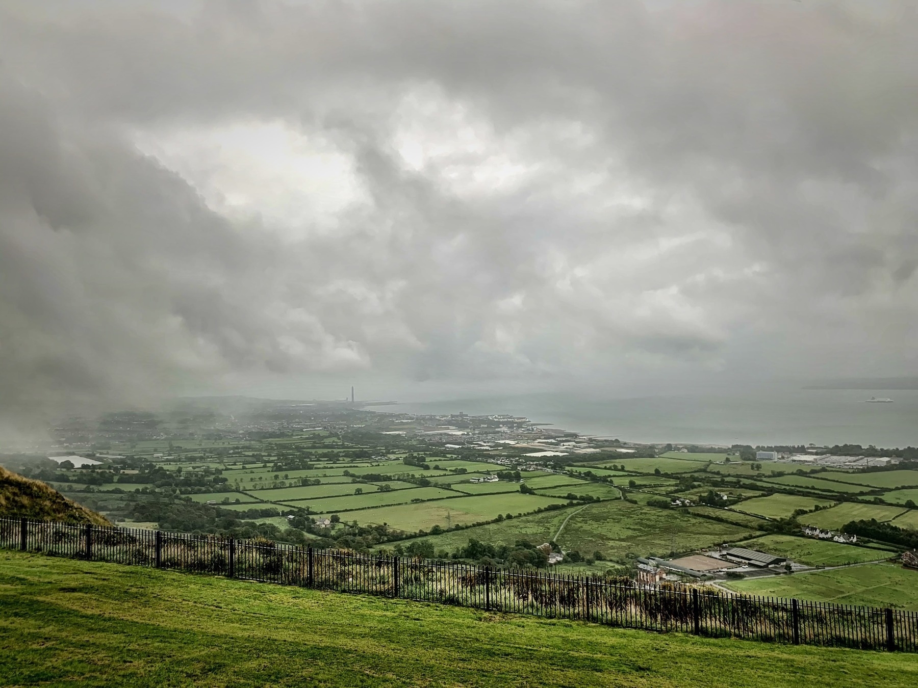 A lush, green landscape with patchwork fields stretches toward a distant body of water under a cloudy, overcast sky.
