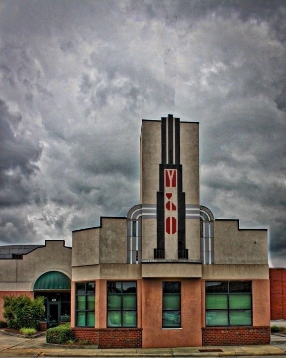Art Deco building stands under stormy skies, featuring vertical and curved architectural lines with red accents. Large green-tinted windows and brick base form the foundation. 