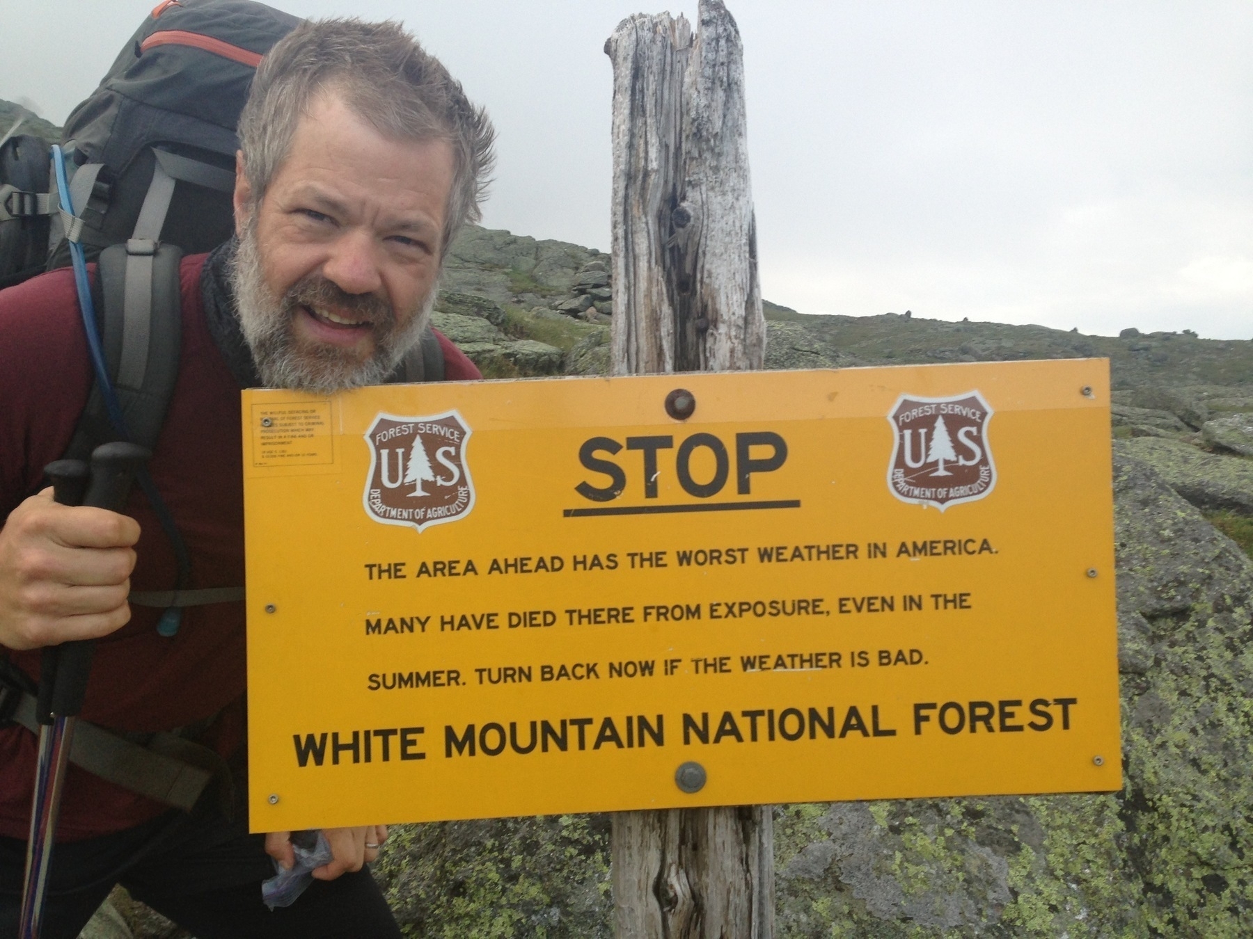 Sign warns hikers, "STOP: The area ahead has the worst weather in America. Many have died from exposure. Turn back if weather is bad. White Mountain National Forest." Hiker stands beside it, outdoors in rocky terrain.