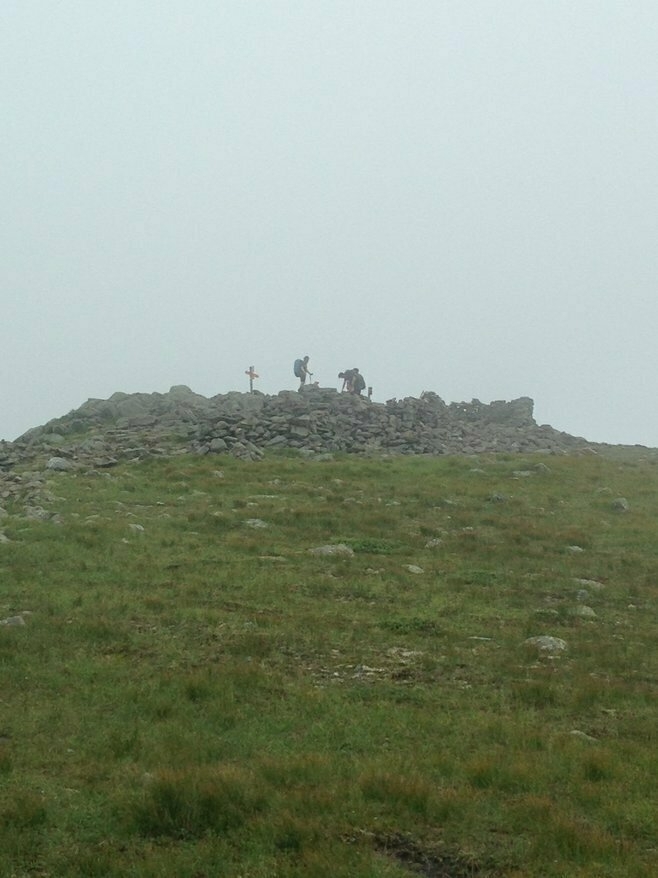 People stand on a rocky summit, adjusting backpacks in a foggy setting. Sparse grass covers the hillside leading up to the peak. A cross or marker is visible at the top.