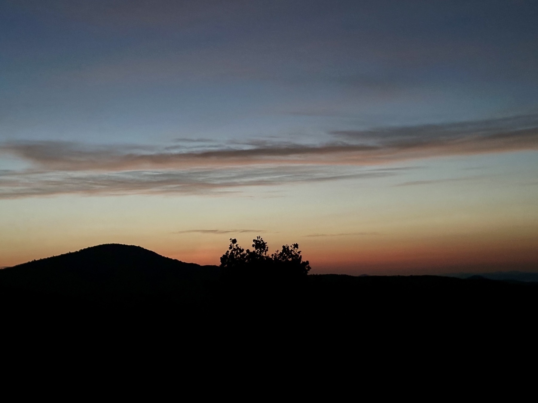 A serene landscape features a silhouetted hill and tree against a colorful dawn sky.