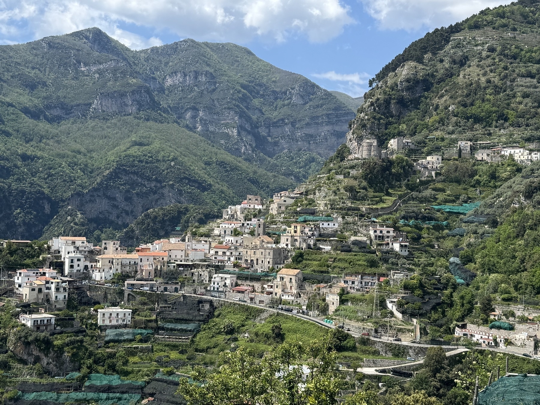 A picturesque hillside village nestled against a backdrop of lush green mountains. The scene features numerous buildings, mostly white with red-tiled roofs, scattered along terraced slopes. Verdant vegetation, including trees and bushes, surrounds the structures, with hints of cultivated fields interspersed throughout. The sky is partly cloudy with patches of blue visible.