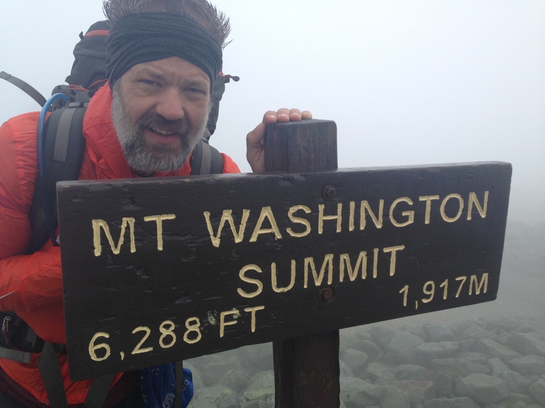 A man stands next to a wooden sign marking the summit of Mt. Washington, which is 6,288 feet or 1,917 meters high.