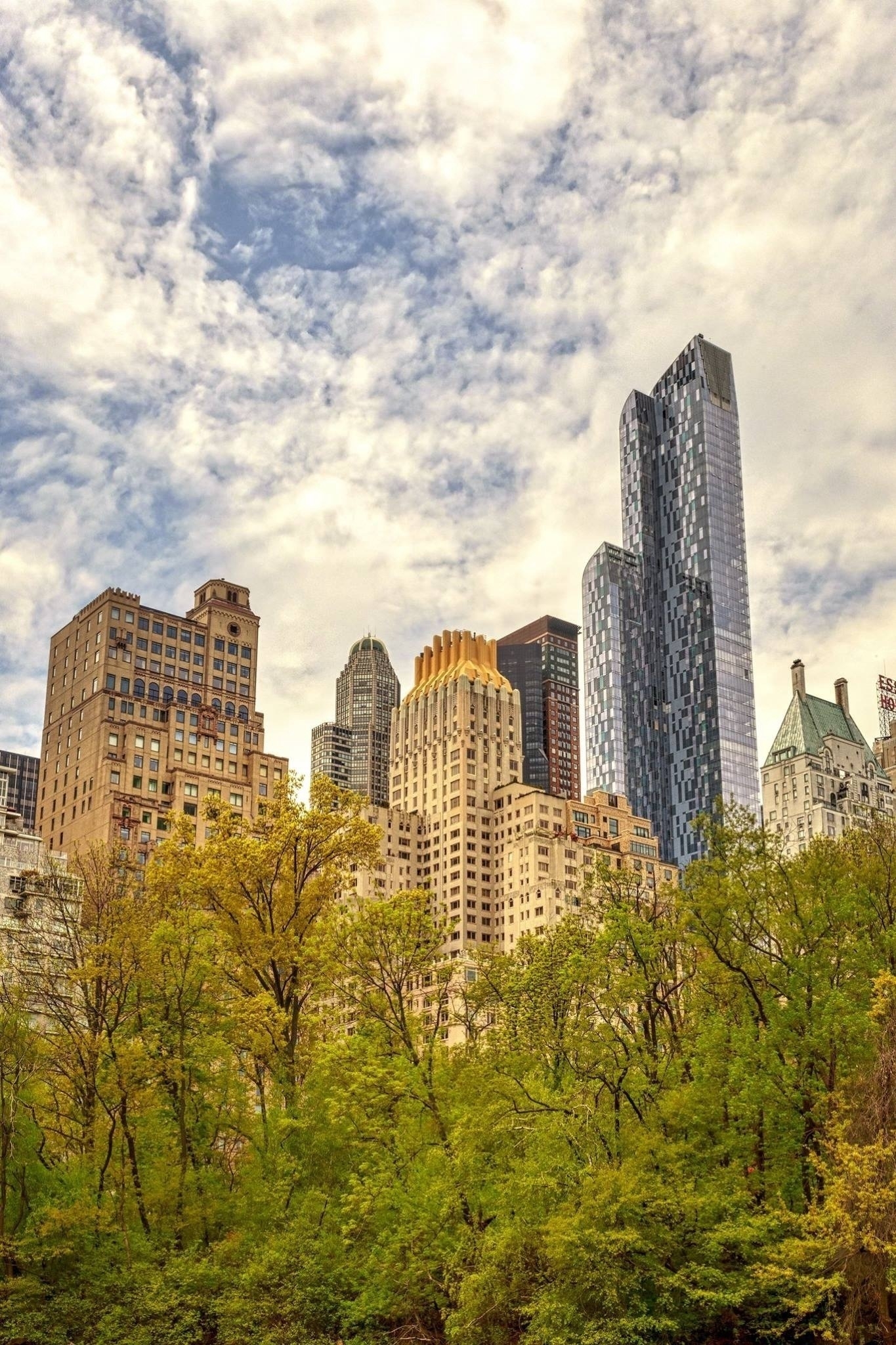 A view of several skyscrapers under a partly cloudy sky. In the foreground, there are lush green trees, contrasting with the tall buildings in the background. The architecture varies, with a mix of modern glass facades and older stone structures.