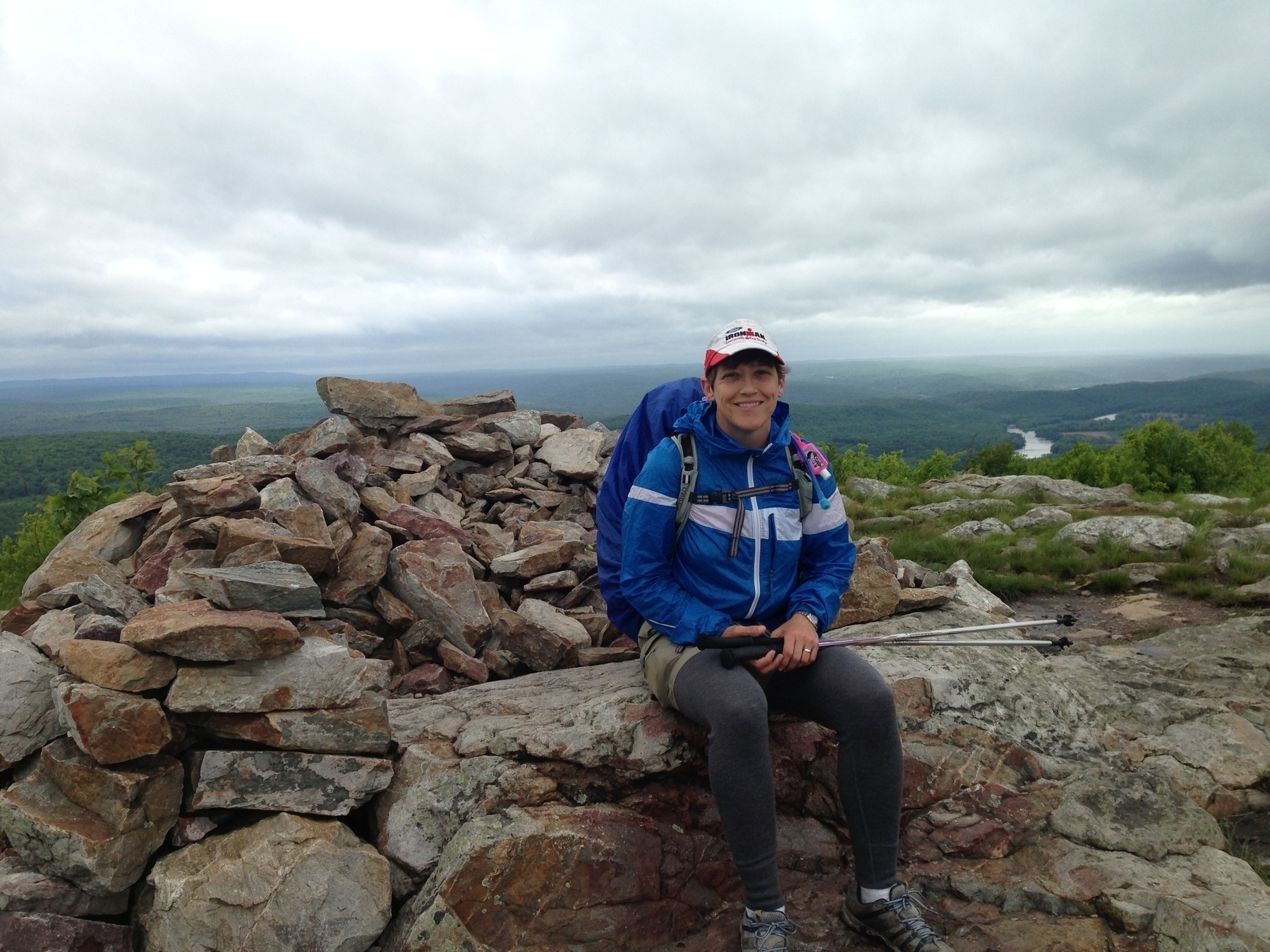 A person wearing hiking gear sits on a rocky mountain summit with a scenic view of the landscape behind them.