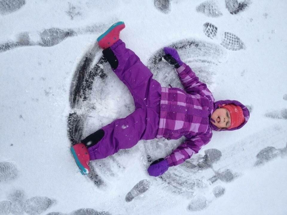 Child in a purple snowsuit makes a snow angel, lying on their back, surrounded by footprints in the snow.