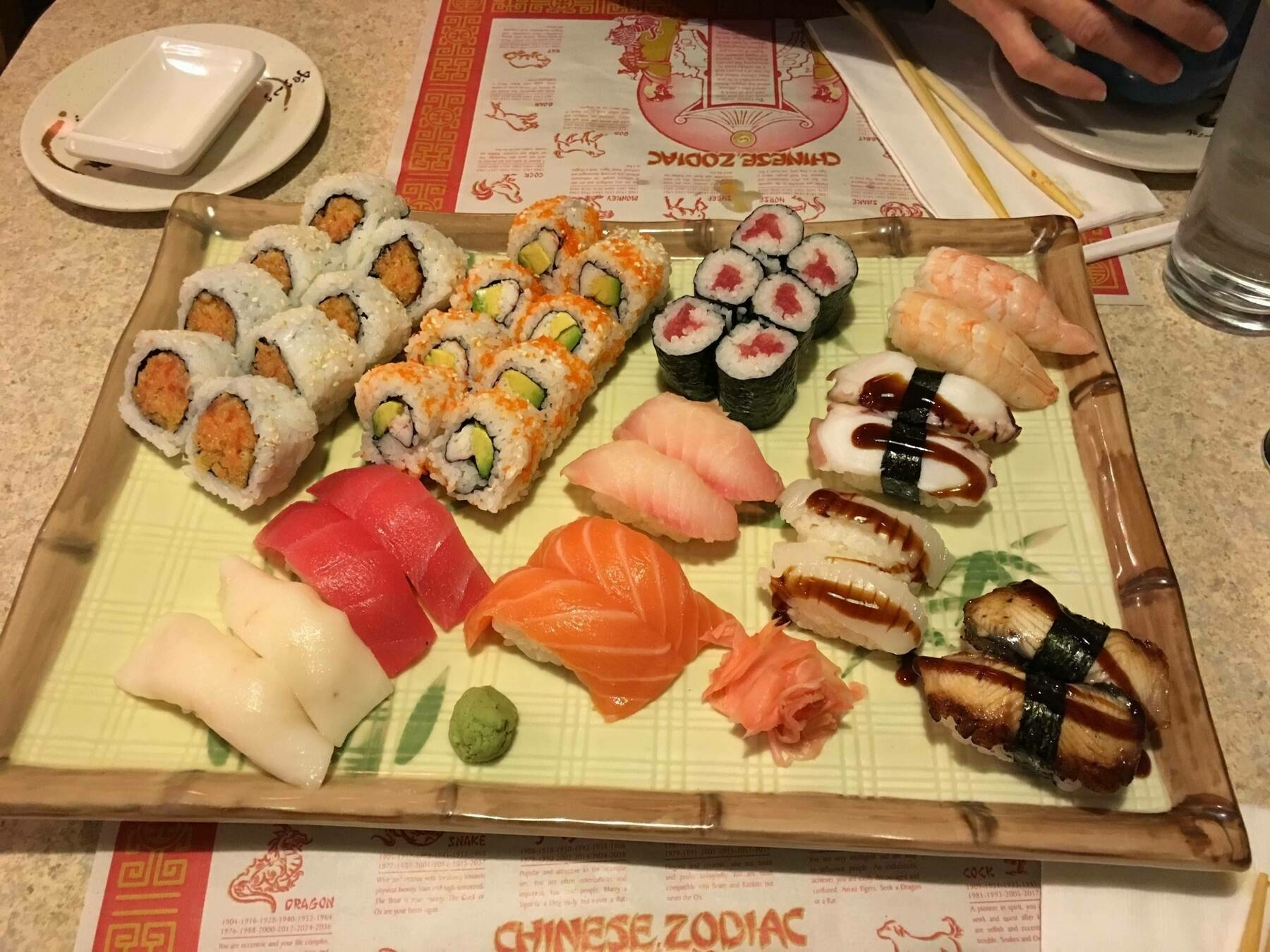 A rectangular plate holds assorted sushi and rolls, including nigiri and maki with various fillings, like tuna and eel, accompanied by wasabi and ginger. Dining table setup nearby.