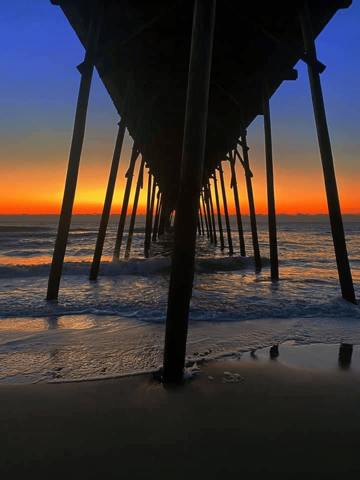 A pier stretches into the sea at sunset, with waves gently washing onto the sandy shore. The sky transitions from deep blue to vibrant orange hues on the horizon.