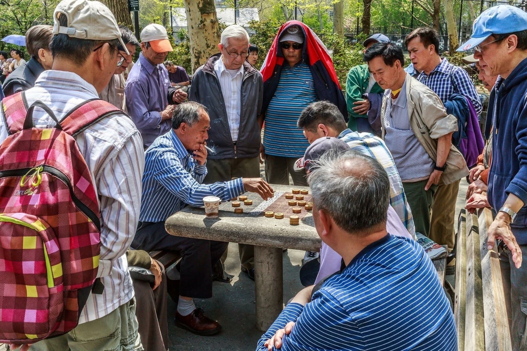 A group of men gather around an outdoor stone table in a park to watch a game of Chinese chess. Two men are seated at the table, deeply engaged in the game. The surrounding spectators, wearing casual clothes and hats, observe attentively. The scene is set on a sunny day with trees and other park-goers in the background.