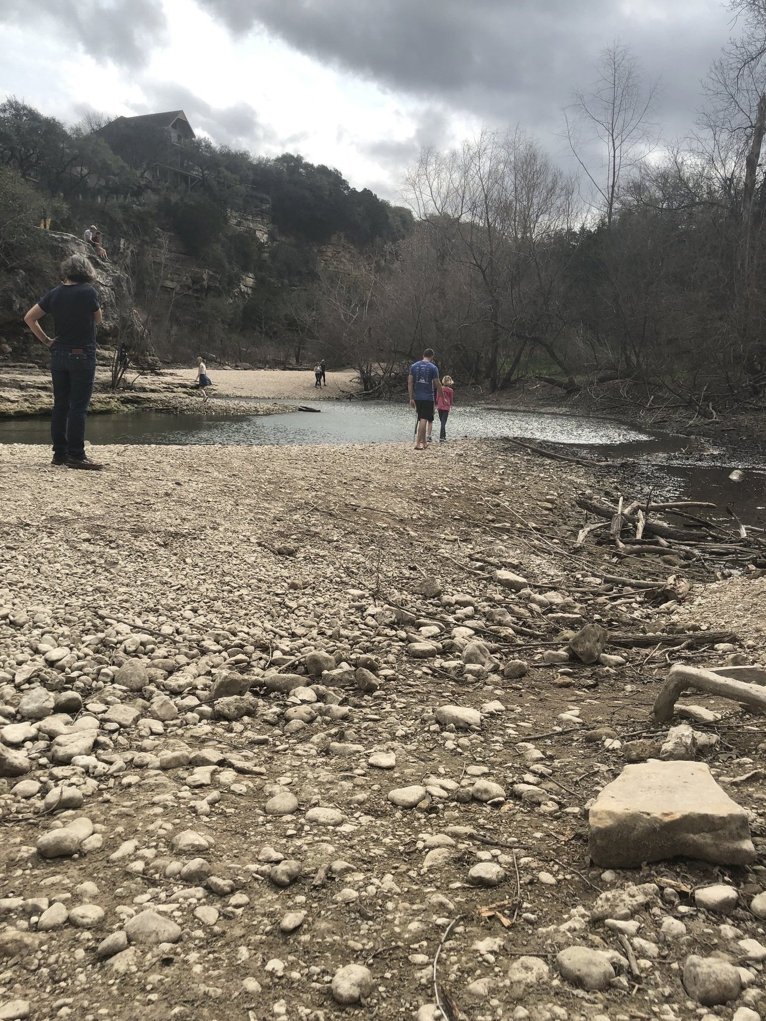 A rocky riverbank with several people scattered around, including a person standing with hands on hips on the left and a man and child walking toward the river on the right. Trees with bare branches line the river, and a cliff with a small house on top is in the background under a cloudy sky.