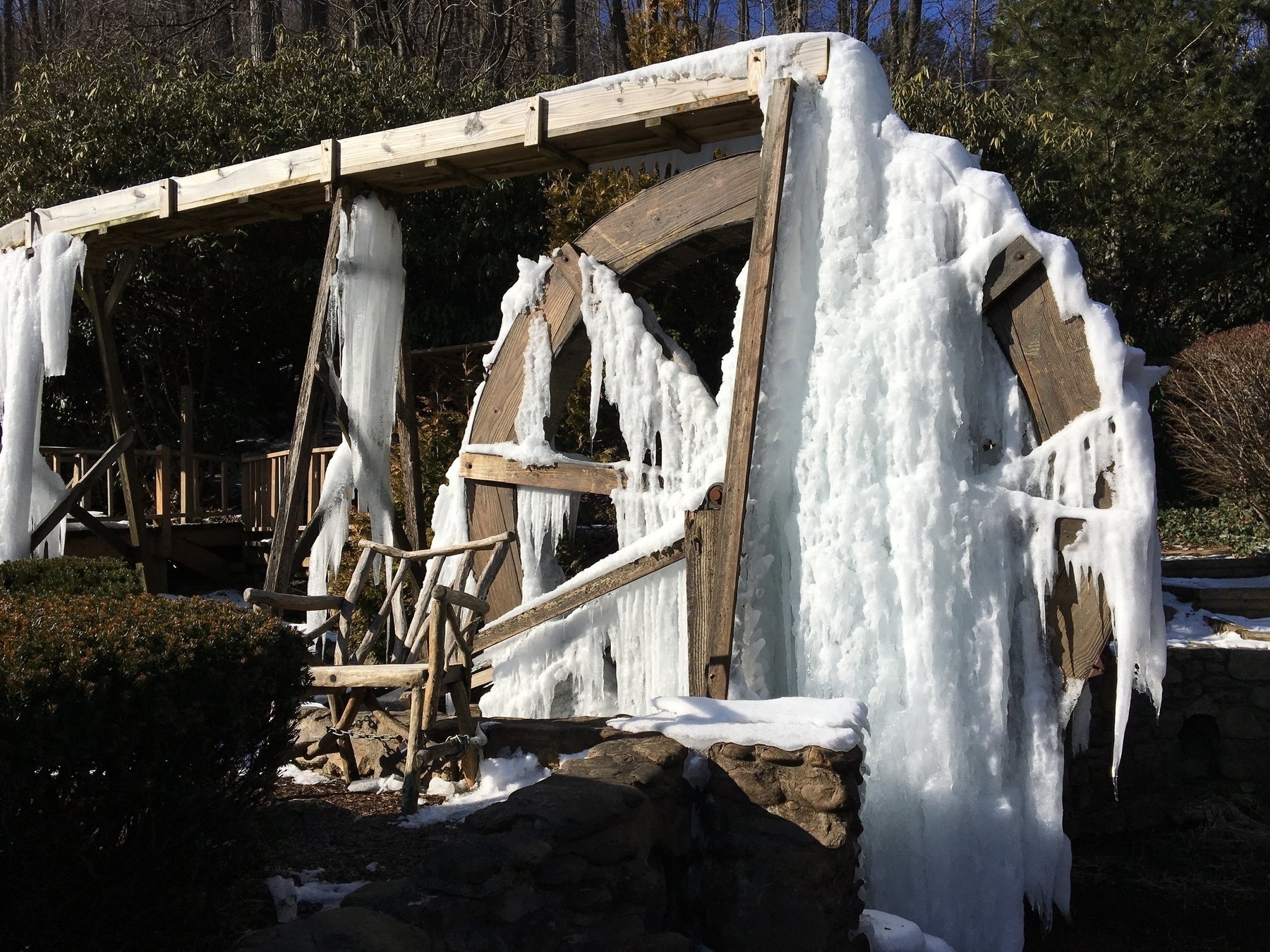 A large wooden waterwheel is covered in thick, cascading icicles, creating a striking winter scene. The structure is set in a garden with surrounding bushes, and the clear blue sky suggests a cold, sunny day. The frozen water adds texture and contrast to the wooden elements of the wheel.