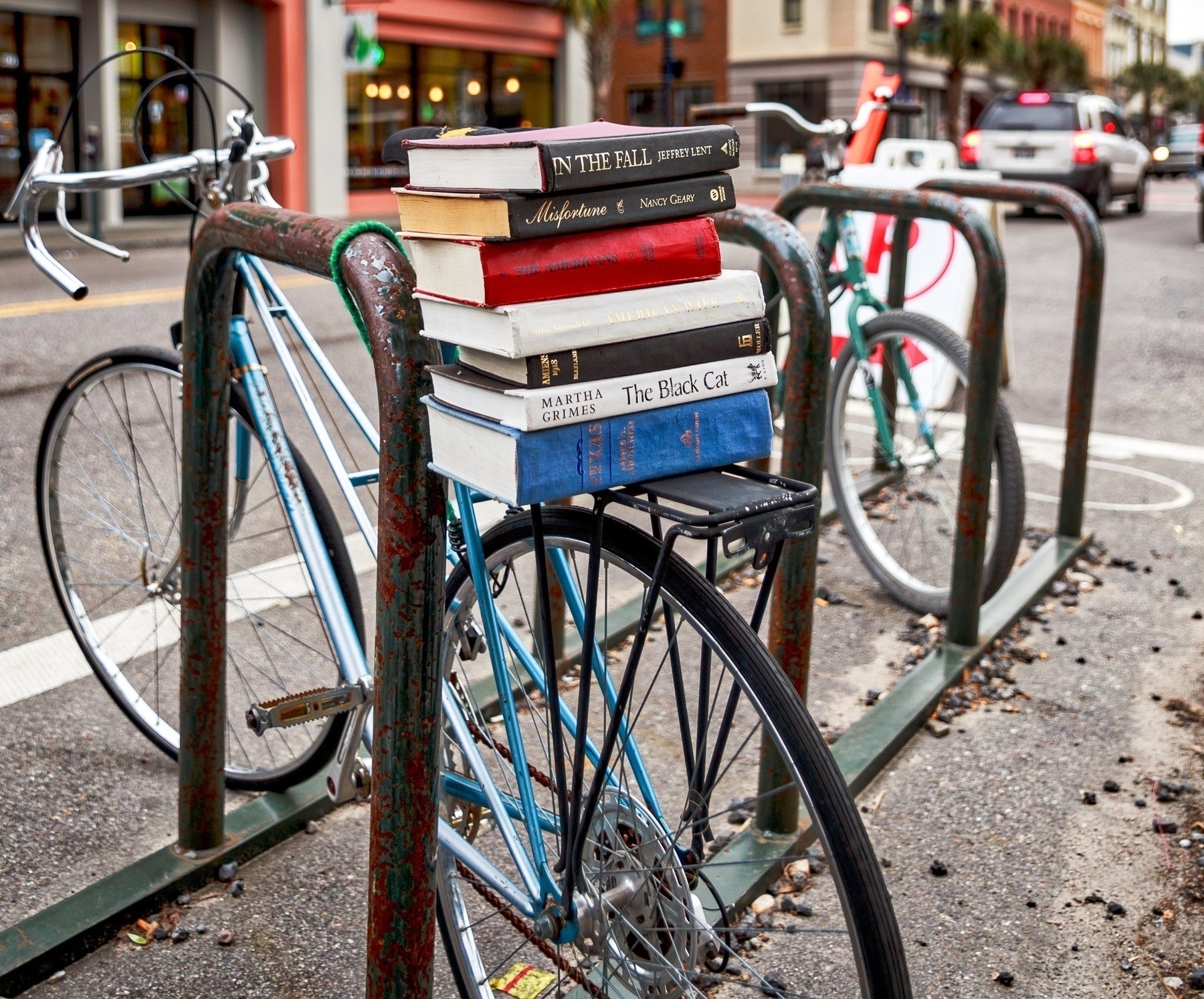 A stack of books is placed on the back rack of a blue bicycle parked in a city street. The rusty metal bike rack holds two bicycles, including the blue one. The street is lined with storefronts and buildings, and cars are visible in the background. The scene suggests an urban setting with a mix of cycling and literary themes.