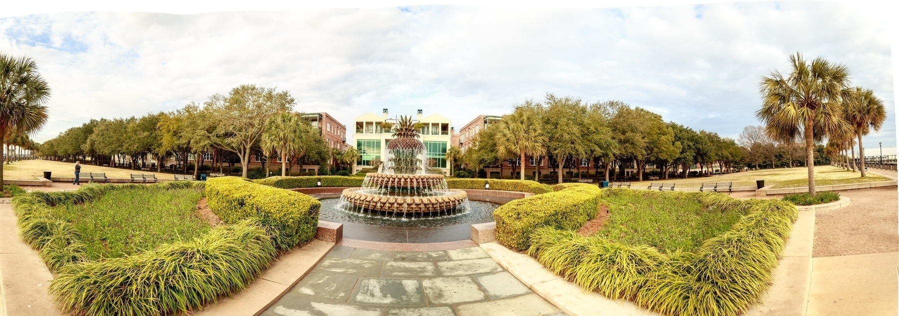 A large fountain shaped like a pineapple is centered in a park. The foreground features manicured hedges and a paved walkway leading to the fountain. On either side of the fountain are palm trees and lush, green lawns. In the background, brick buildings and more trees are visible under a cloudy sky.