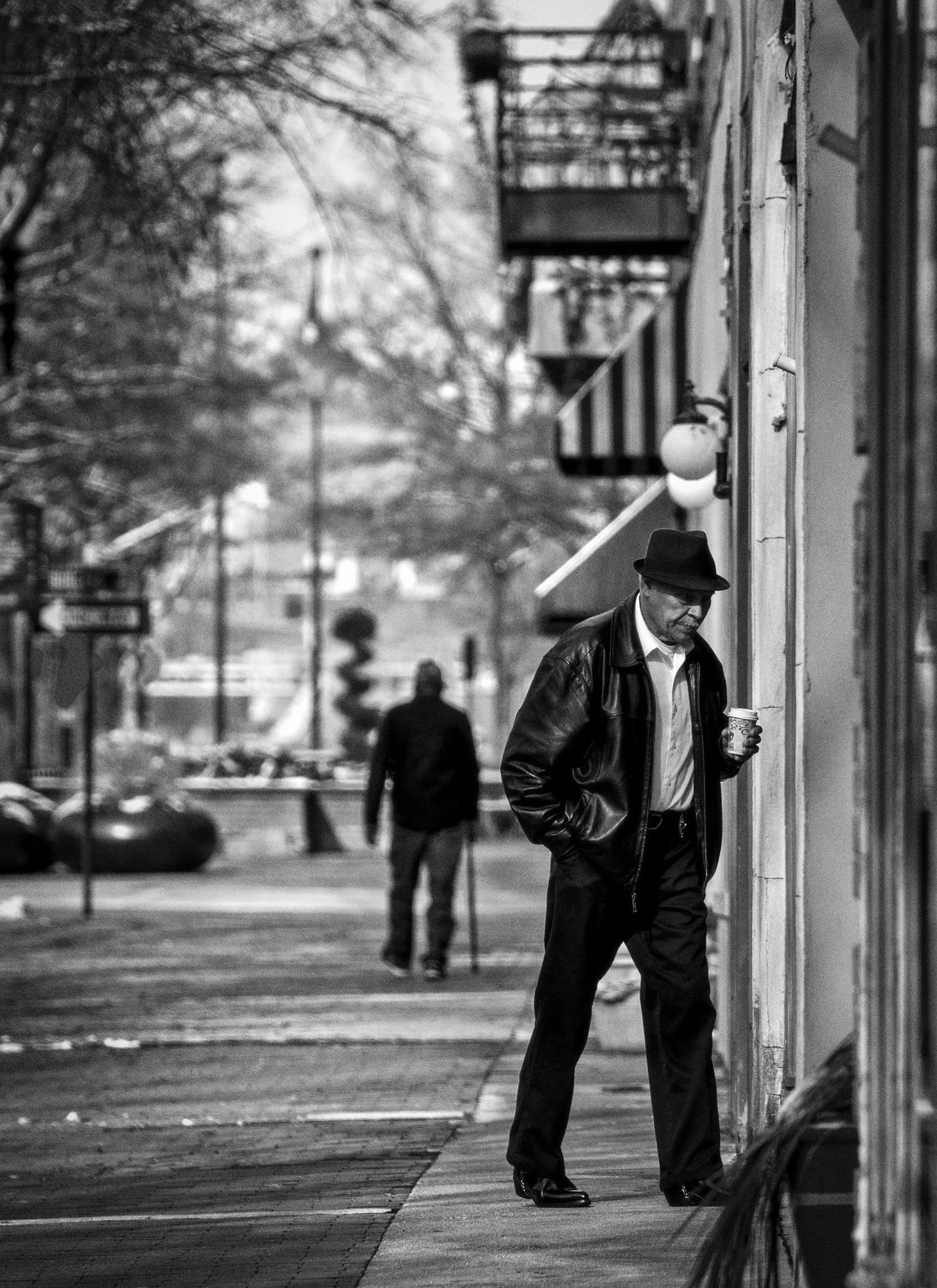 Black and white image of a man walking along a city sidewalk, wearing a fedora hat, leather jacket, and holding a coffee cup. He is near a building wall with spherical light fixtures. In the background, another person is walking away, trees and street elements are visible.