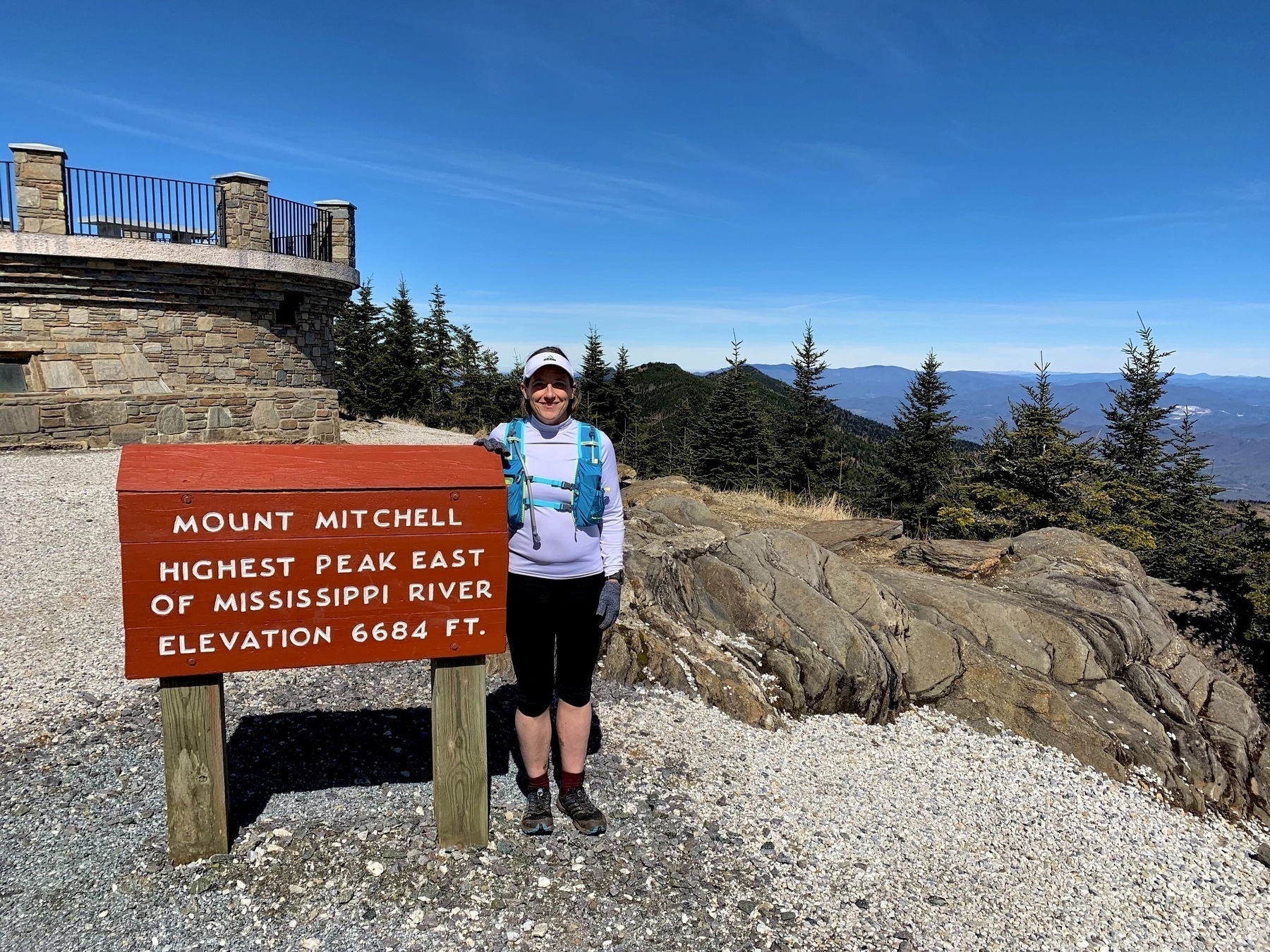 A person stands beside a sign indicating the elevation of Mount Mitchell, with a stone structure and scenic mountainous landscape in the background.