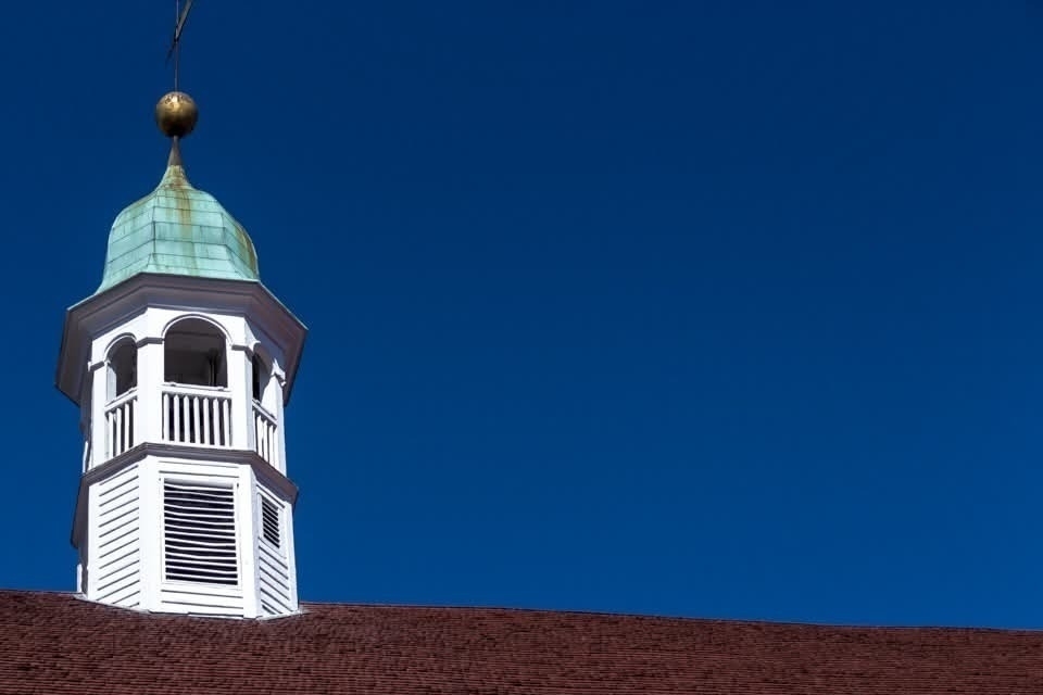 A white cupola with a copper-green roof and a golden ball on top extends above a dark red roof against a clear blue sky. The cupola features slatted windows and railing details.