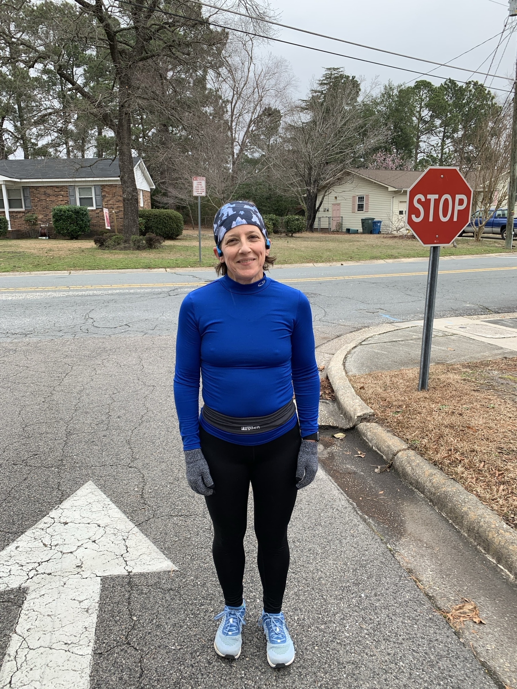 A person stands smiling on a road wearing a blue shirt, black leggings, and gloves. Nearby is a stop sign and residential houses with trees in the background.