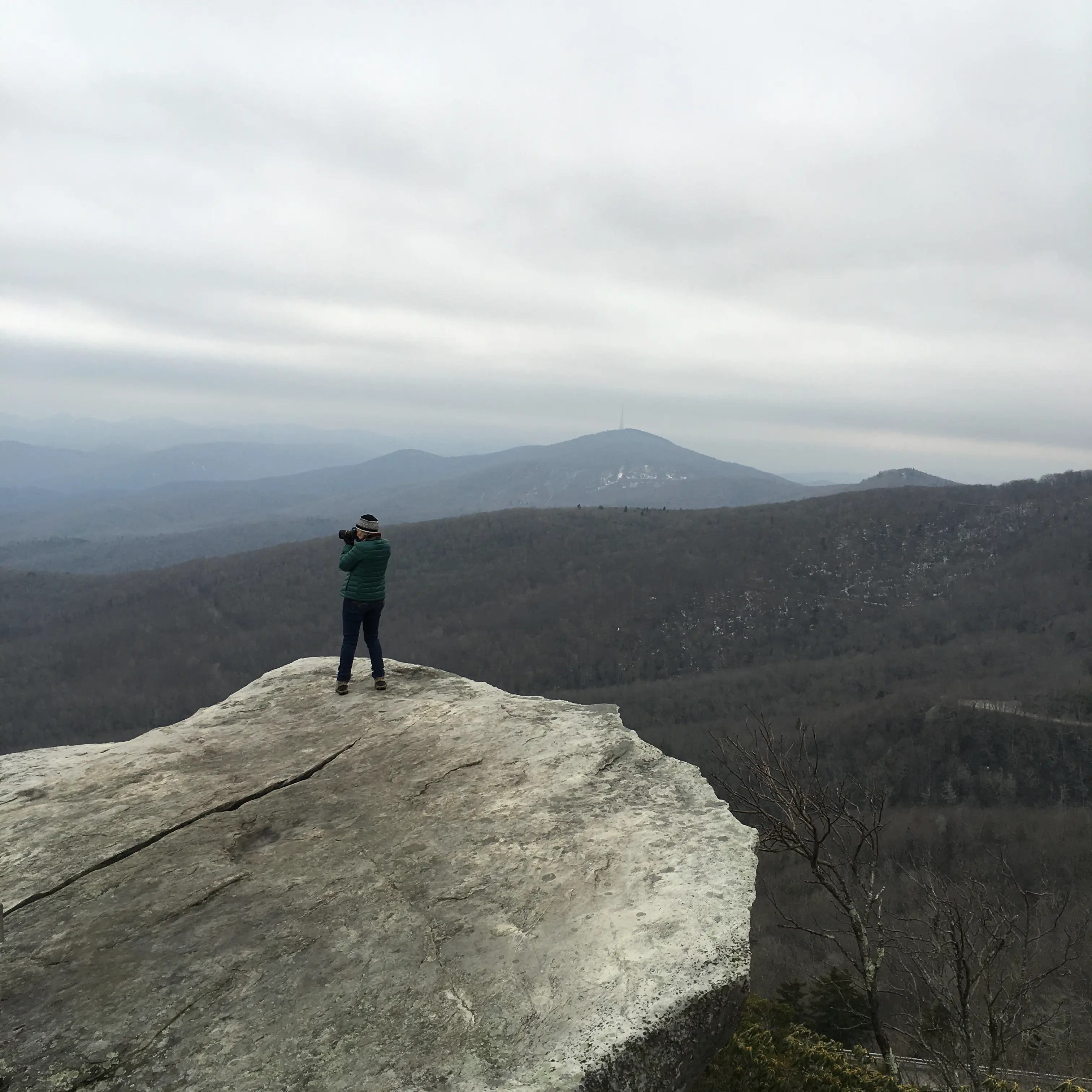 A person stands photographing on a large, flat rock overlooking rolling, forested hills under a cloudy sky.